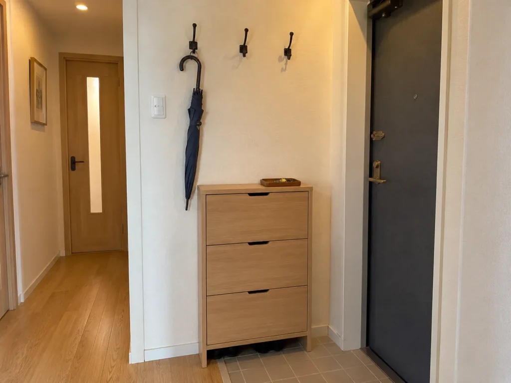 Light wood tilt-out shoe cabinet next to a dark front door, with wall-mounted hooks holding an umbrella and a view of an adjacent hallway.